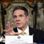 Andrew Cuomo speaking at a press conference, wearing a suit.