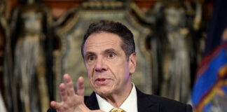 Andrew Cuomo speaking at a press conference, wearing a suit.