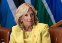Jill Biden in a yellow blazer sitting, colorful flags behind.