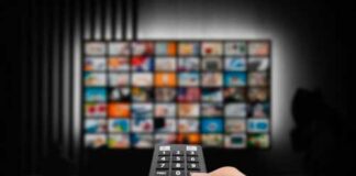 Hand holding a remote control in front of a television displaying various media options