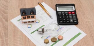 A miniature house model, light bulb, coins, calculator, and financial documents on a wooden surface