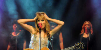 Taylor Swift holding guitar on stage, background singers behind.