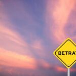 A yellow warning sign with the word 'BETRAY' against a colorful sunset sky