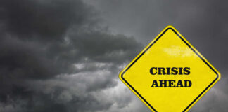 A yellow warning sign that reads 'CRISIS AHEAD' against a backdrop of dark, stormy clouds
