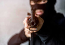 Person in balaclava aiming rifle at the camera.