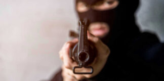 Person in balaclava aiming rifle at the camera.