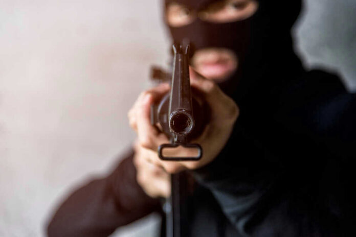 Person in balaclava aiming rifle at the camera.