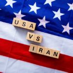 A wrecking ball painted with the American flag is breaking through a wall featuring the Iranian flag