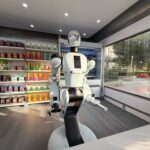 A humanoid robot using a tablet in an industrial setting with robotic arms in action