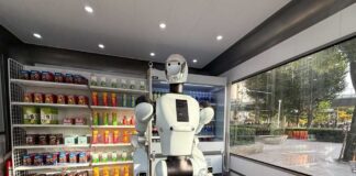 A humanoid robot using a tablet in an industrial setting with robotic arms in action