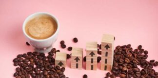 A cup of coffee next to wooden blocks with upward arrows and scattered coffee beans on a pink background