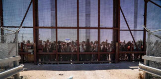 Father Dies In ICE Custody People standing behind a fence on a dusty path.