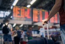 A traveler holding a suitcase in an airport terminal with a flight information display in the background
