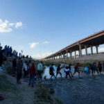 People crossing a shallow river near an overpass